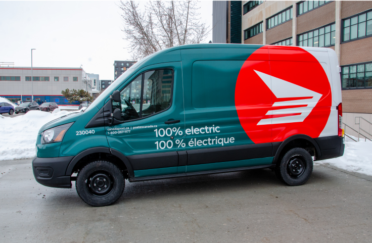 Un véhicule de livraison électrique écologique de Postes Canada est stationné à l’extérieur d’une installation de l’entreprise. On peut lire « 100 % électrique » sur le côté. Il est vert forêt et blanc, et porte l’icône du logo rouge de Postes Canada
