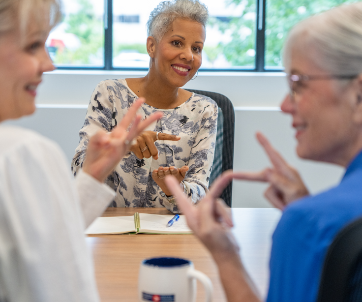 Une femme aux cheveux gris parle en langage des signes avec une femme blonde et une autre aux cheveux gris dans une salle de conférence